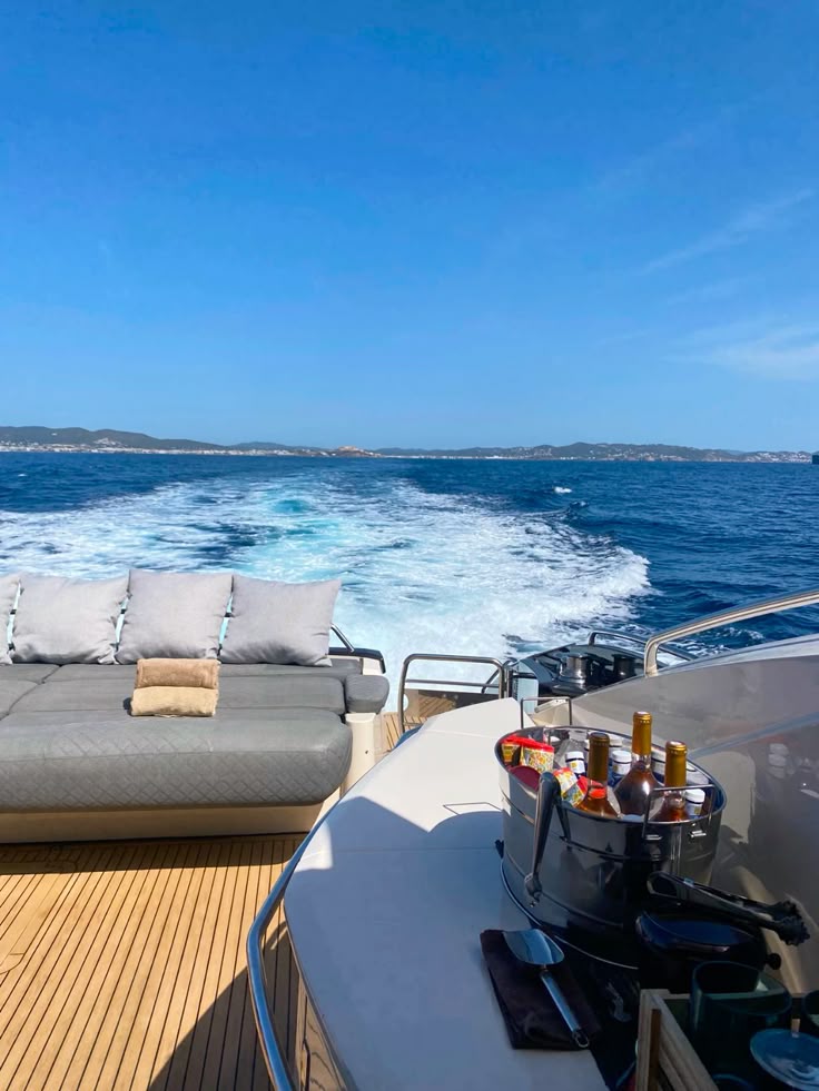 View from the back of a luxury yacht cruising on a sunny day, with a cushioned lounge area, an ice bucket with drinks, and a foamy wake leading to the blue sea and distant coastline.
