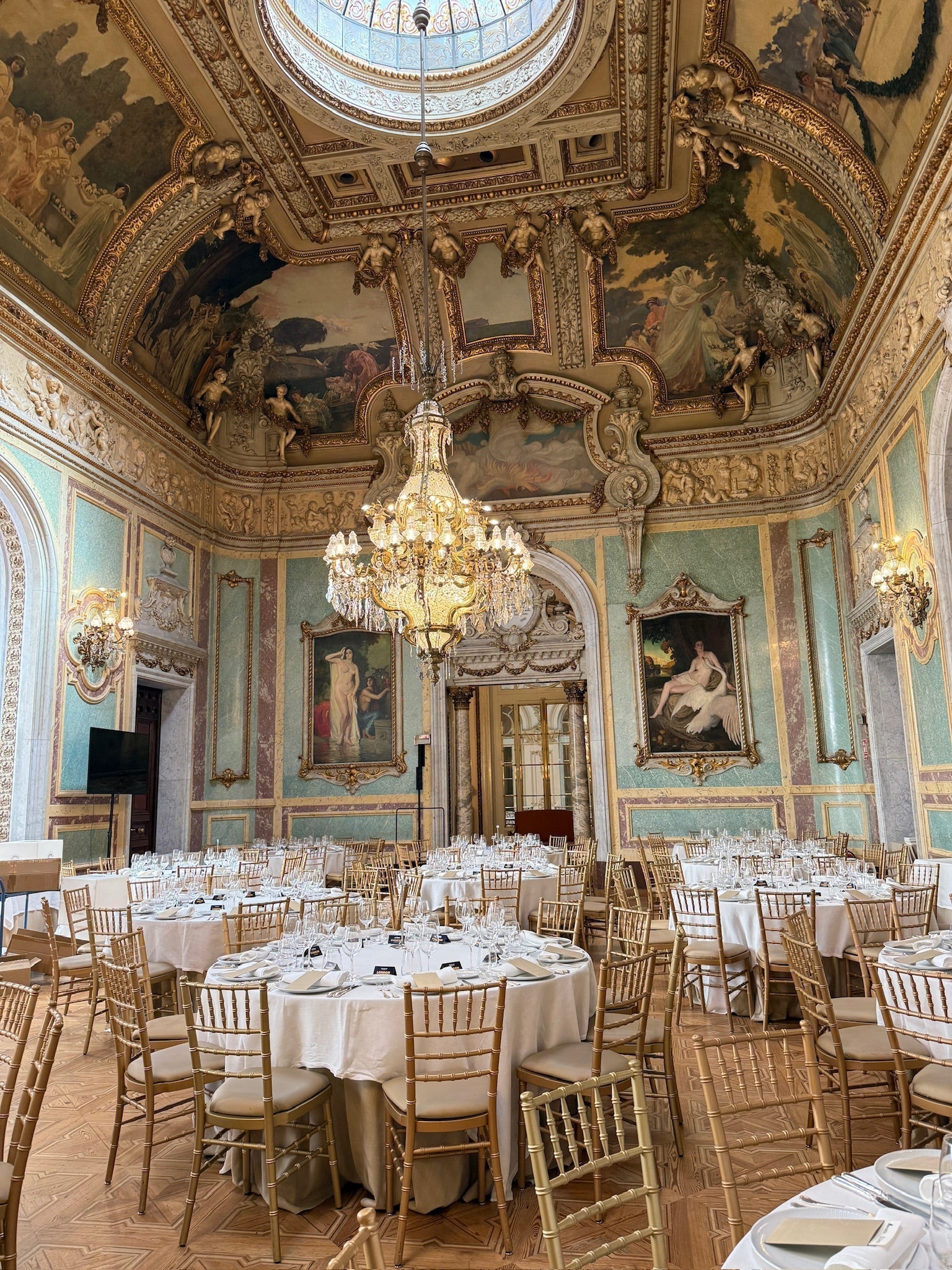 Elegant corporate event setup in a historic Barcelona venue featuring ornate ceilings, crystal chandeliers, round banquet tables, and gold Chiavari chairs, creating a luxurious atmosphere for business events and gala dinners.