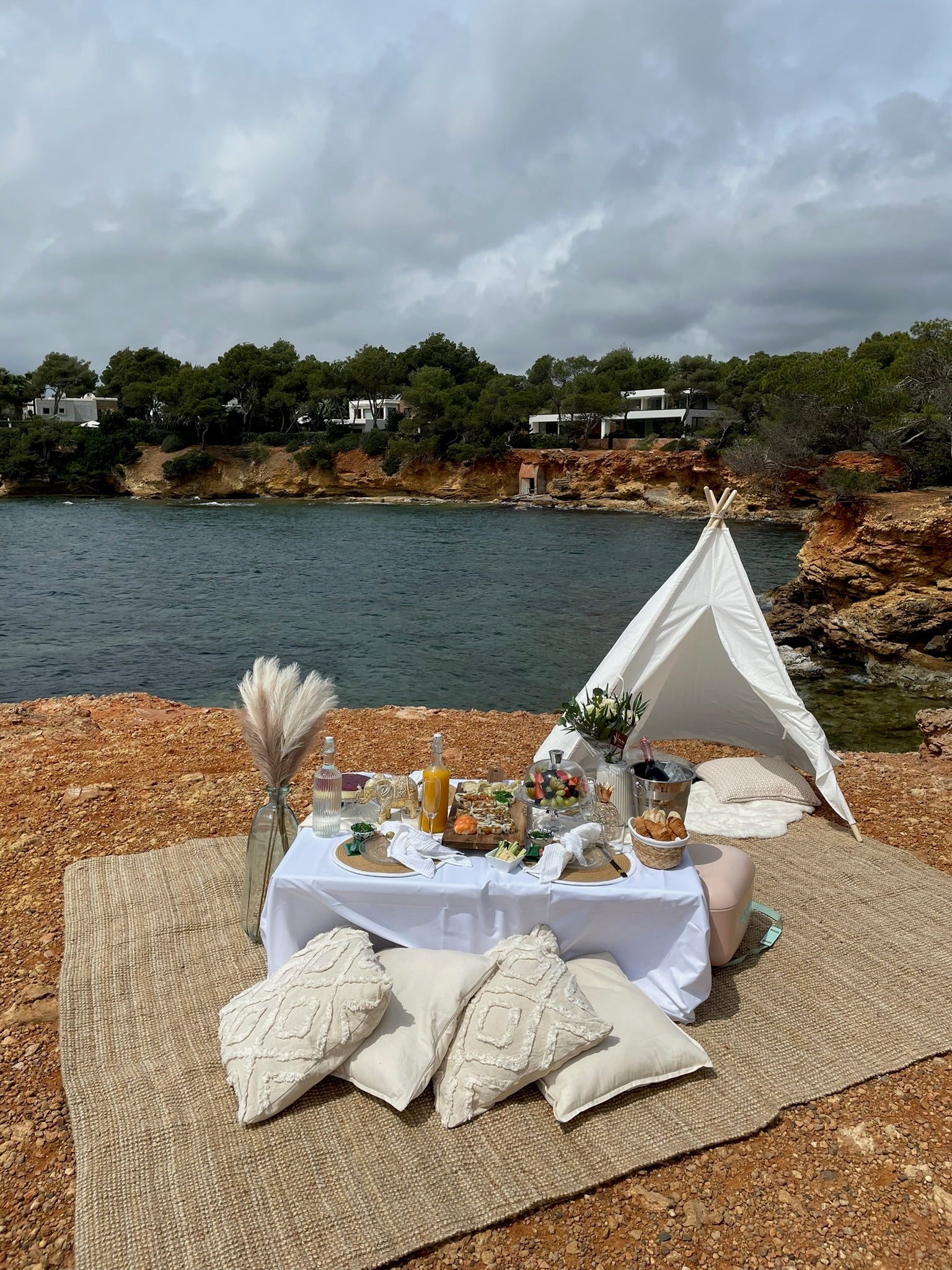 Luxury picnic setup on a coastal cliff in Mallorca with a low table, cushions, teepee tent, and food, overlooking the sea.
