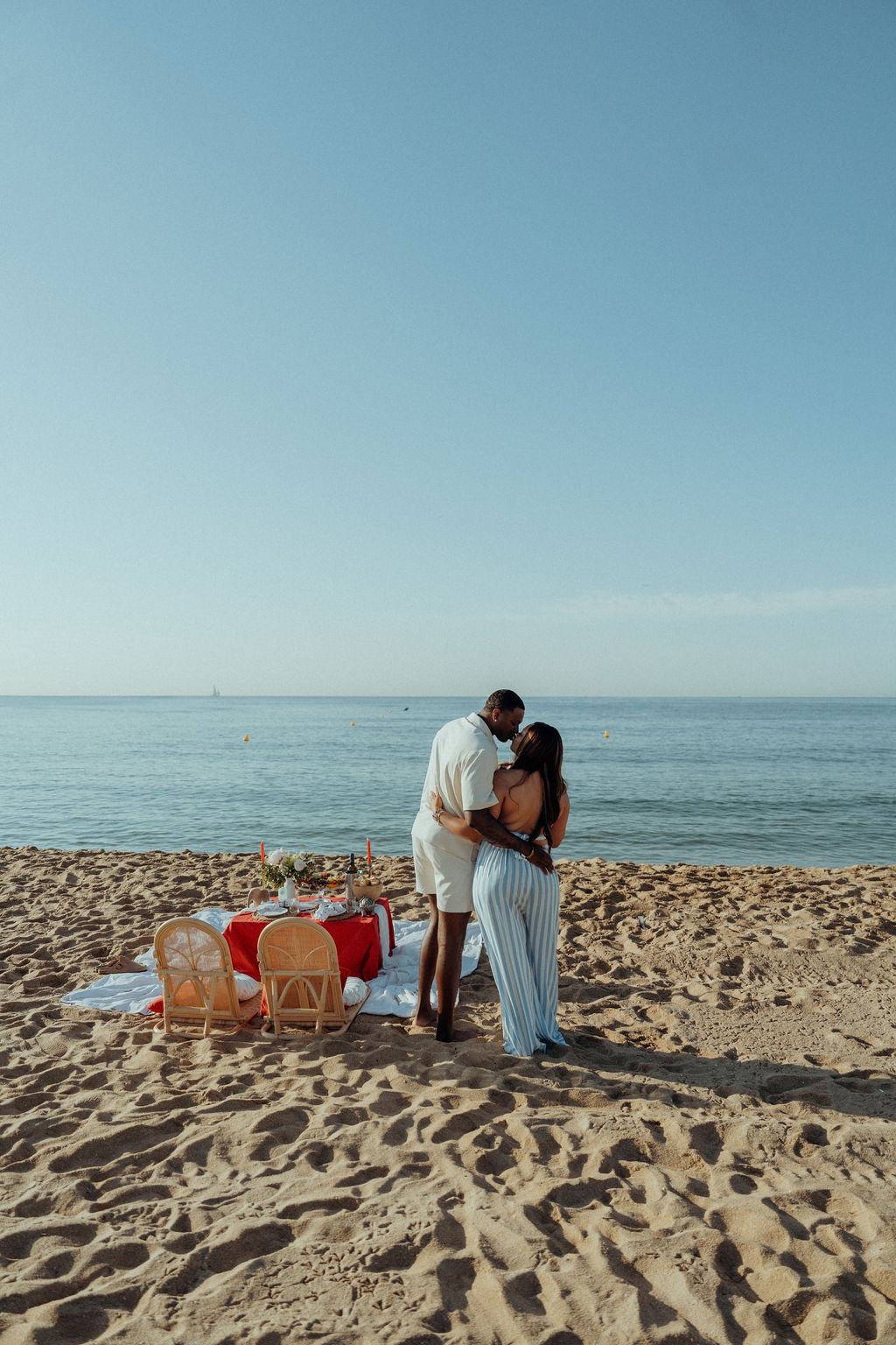Romantic beach picnic setup with a couple embracing by the sea, featuring a styled low table with candles, flowers, grazing food and drinks, creating an intimate proposal or date experience at sunset.