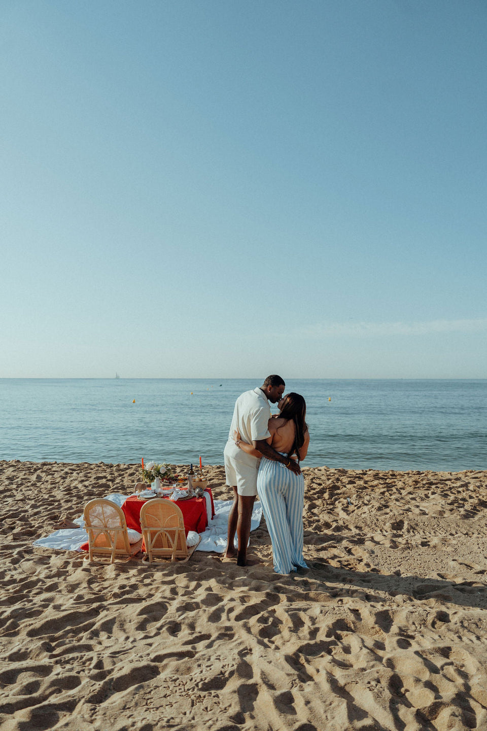 Romantic beach picnic setup with a couple embracing by the sea, featuring a styled low table with candles, flowers, grazing food and drinks, creating an intimate proposal or date experience at sunset.