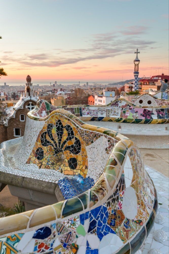 Colourful Gaudí mosaic terrace in Park Güell overlooking Barcelona at sunset, with pastel sky and city skyline in the background.