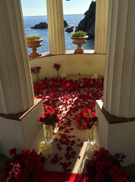 Romantic seaside proposal setup with red rose petals, roses, and candles framed by elegant columns overlooking the ocean.