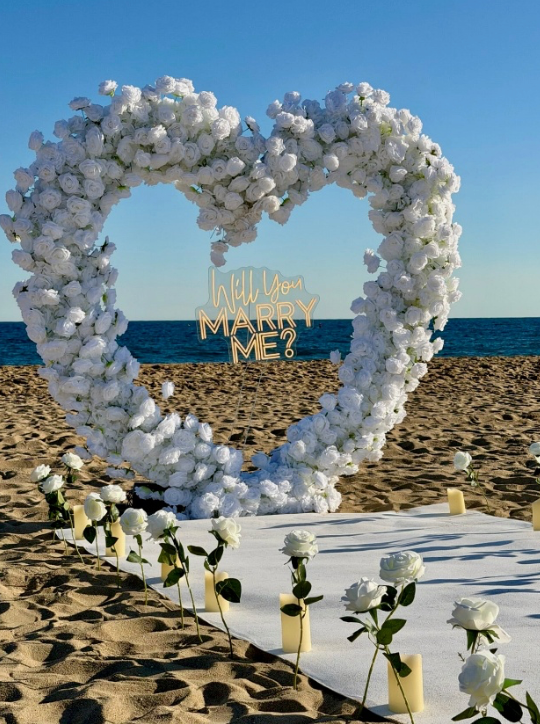Romantic beach proposal setup featuring a heart-shaped white floral arch with a “Will You Marry Me?” sign, candle-lined aisle, and white roses on the sand, overlooking the sea under a clear blue sky.