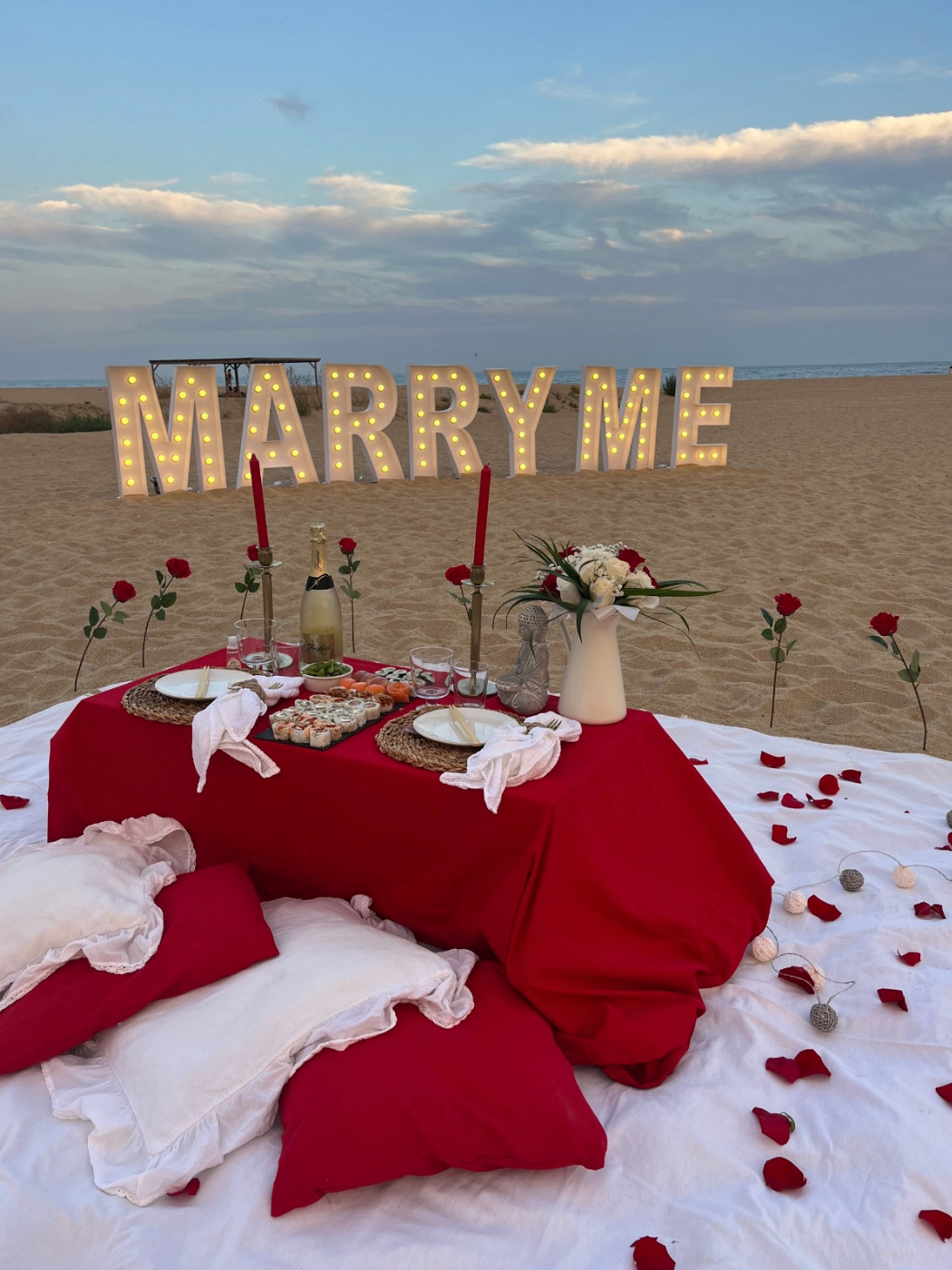 Romantic beach proposal picnic with a red-themed setup, cozy floor cushions, candlelit table, roses, champagne, and illuminated “MARRY ME” letters on the sand, creating an intimate and unforgettable seaside proposal moment.