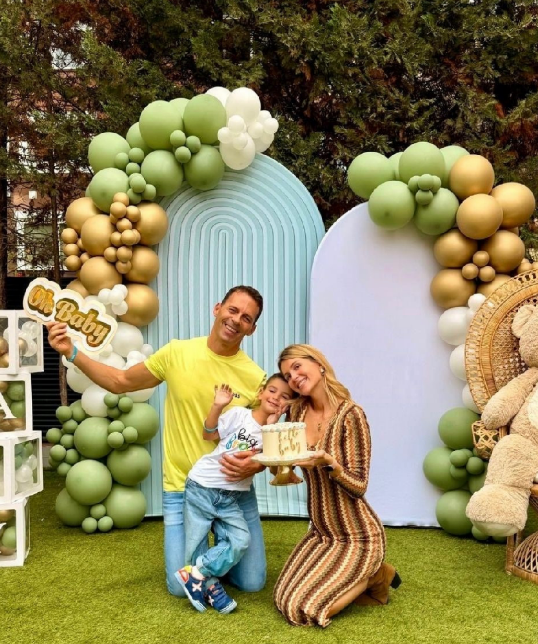 Family celebrating at a stylish “Oh Baby” outdoor setup with pastel blue and white backdrops, green and gold balloon arches, and a cozy teddy-bear corner—perfect for a baby shower or gender reveal photo moment.
