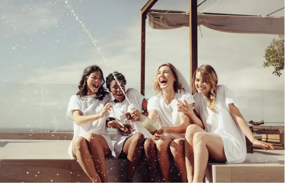 Group of bridesmaids in white outfits laughing and spraying champagne during a fun hen party celebration on a sunny terrace in Barcelona, organised by Straws ’n Berries.