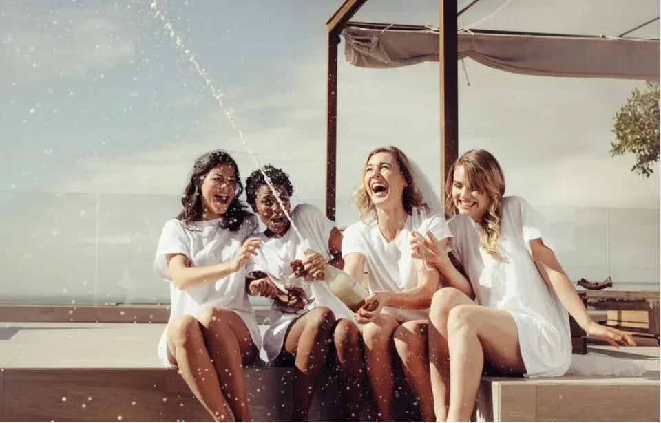 Group of bridesmaids in white outfits laughing and spraying champagne during a fun hen party celebration on a sunny terrace in Barcelona, organised by Straws ’n Berries.