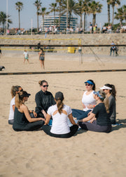 A group of seven people sitting in a circle on a sandy beach volleyball court, with palm trees and a building in the background.