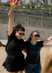 Two women playing beach volleyball on a sunny day, with one holding up a pink and green paddle and both wearing black athletic outfits and sunglasses.