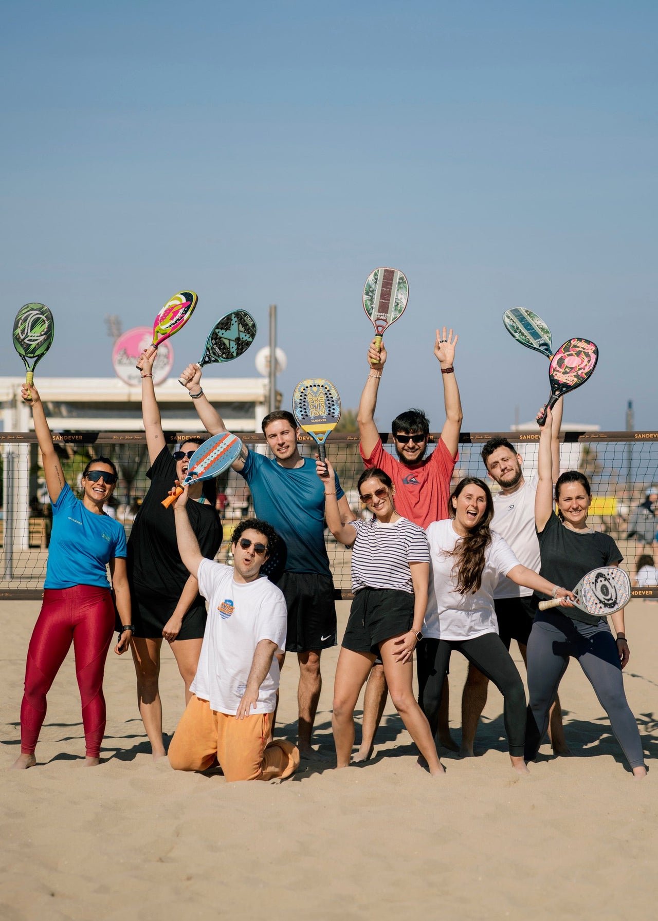 A group of nine people posing together on a sandy beach, each holding up a different colored paddle racket above the net. The group is dressed in casual athletic wear, and the background shows a clear blue sky and the ocean.