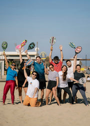A group of nine people posing together on a sandy beach, each holding up a different colored paddle racket above the net. The group is dressed in casual athletic wear, and the background shows a clear blue sky and the ocean.
