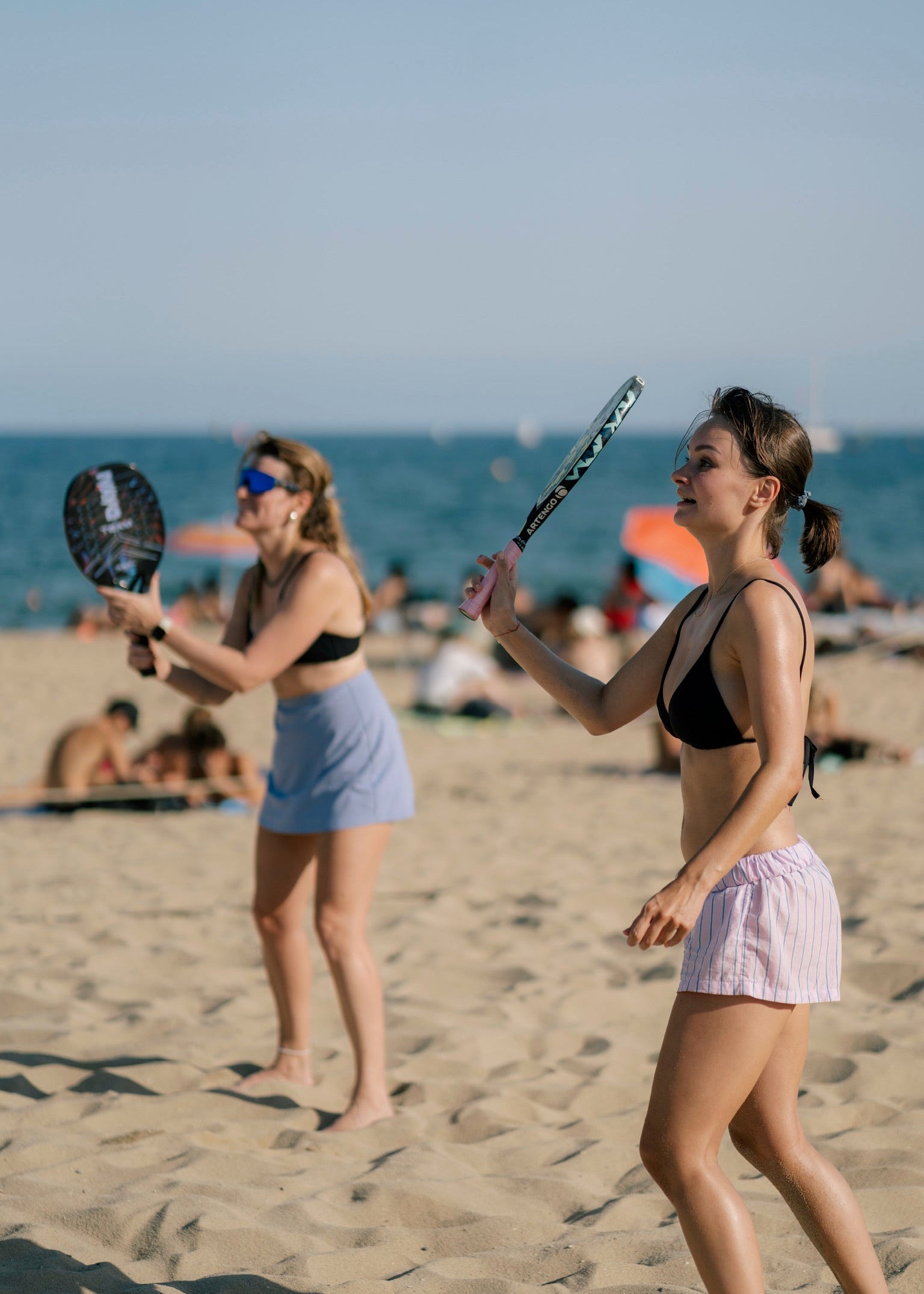 Two women playing paddle tennis on a sandy beach with the ocean and sky in the background.