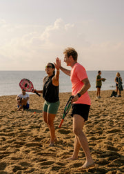 Two people high-fiving on a sandy beach while playing paddle tennis, with the ocean and other beachgoers in the background.
