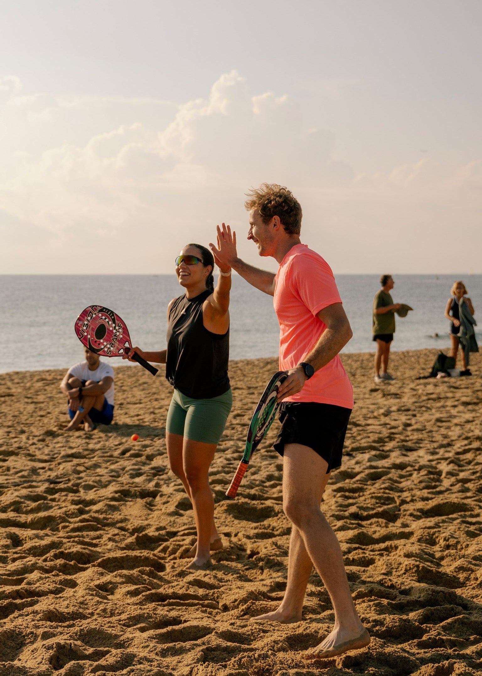 Two people high-fiving on a sandy beach while playing paddle tennis, with the ocean and other beachgoers in the background.