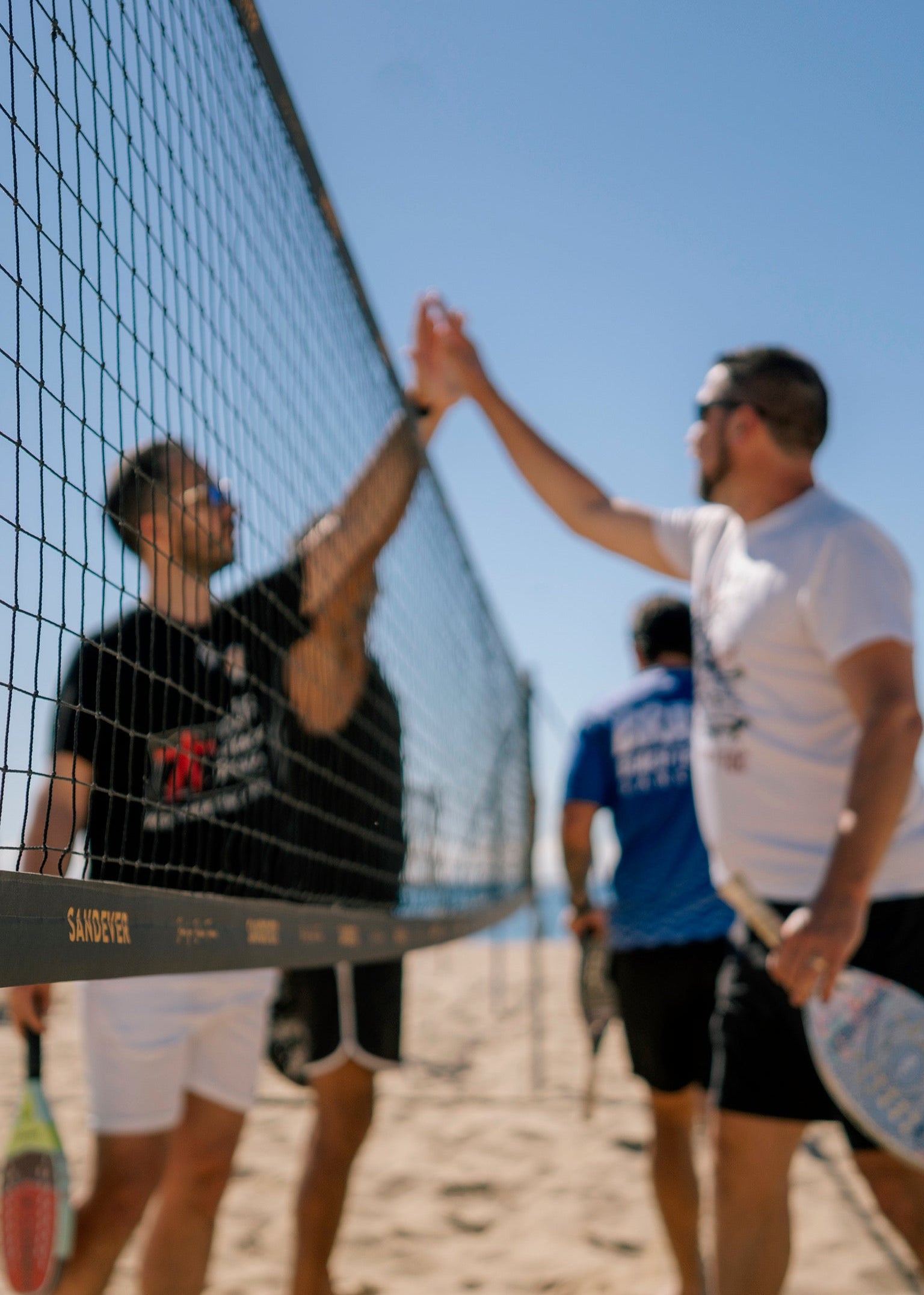 Three men playing beach volleyball on a sandy court, with two players reaching to high-five over the net. One player in a white shirt and black shorts is in sharp focus in the foreground, while the other players are blurred in the background under a clear blue sky.
