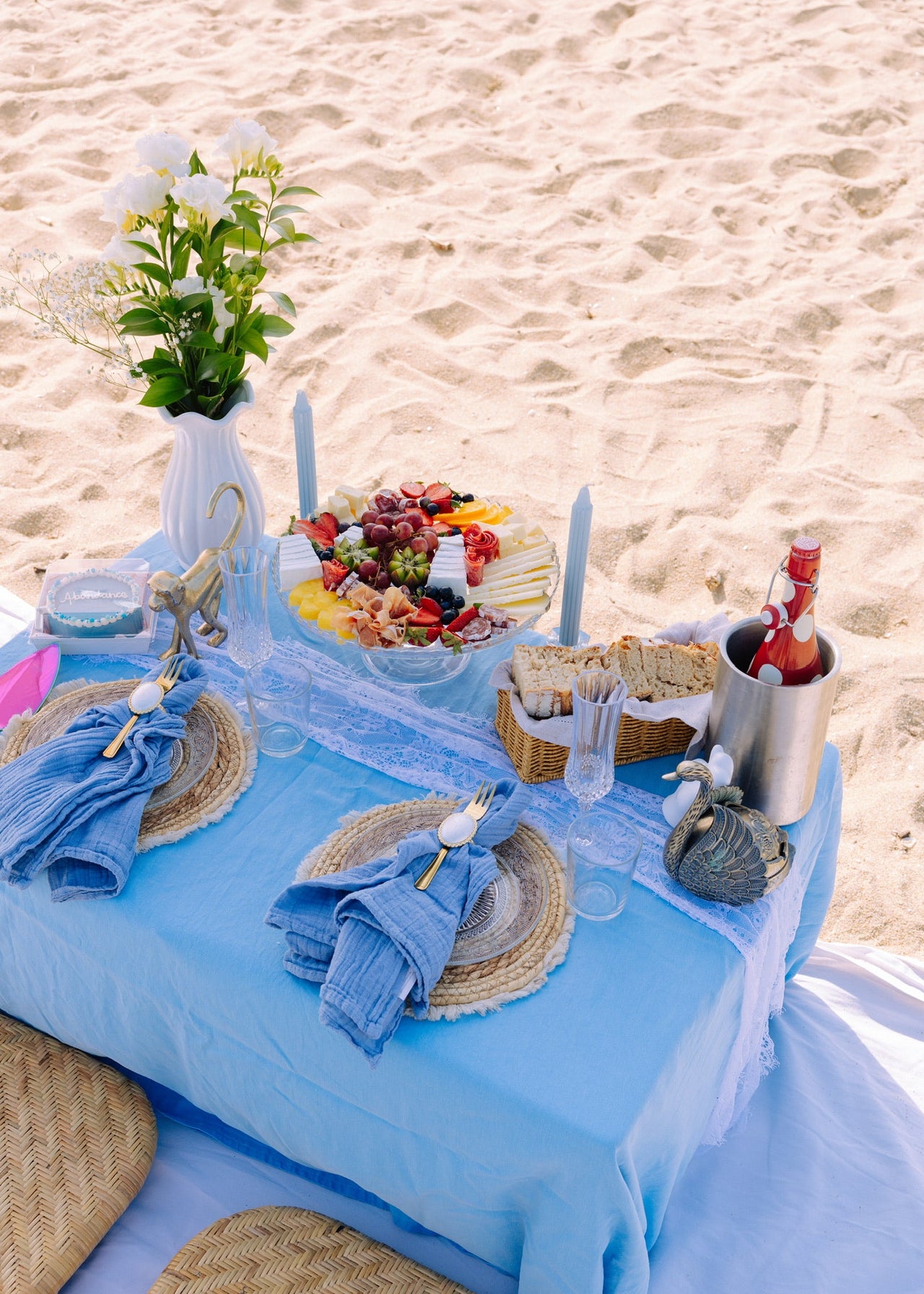 Beachside table setting with blue tablecloth, fruit platter, and candles on sandy background