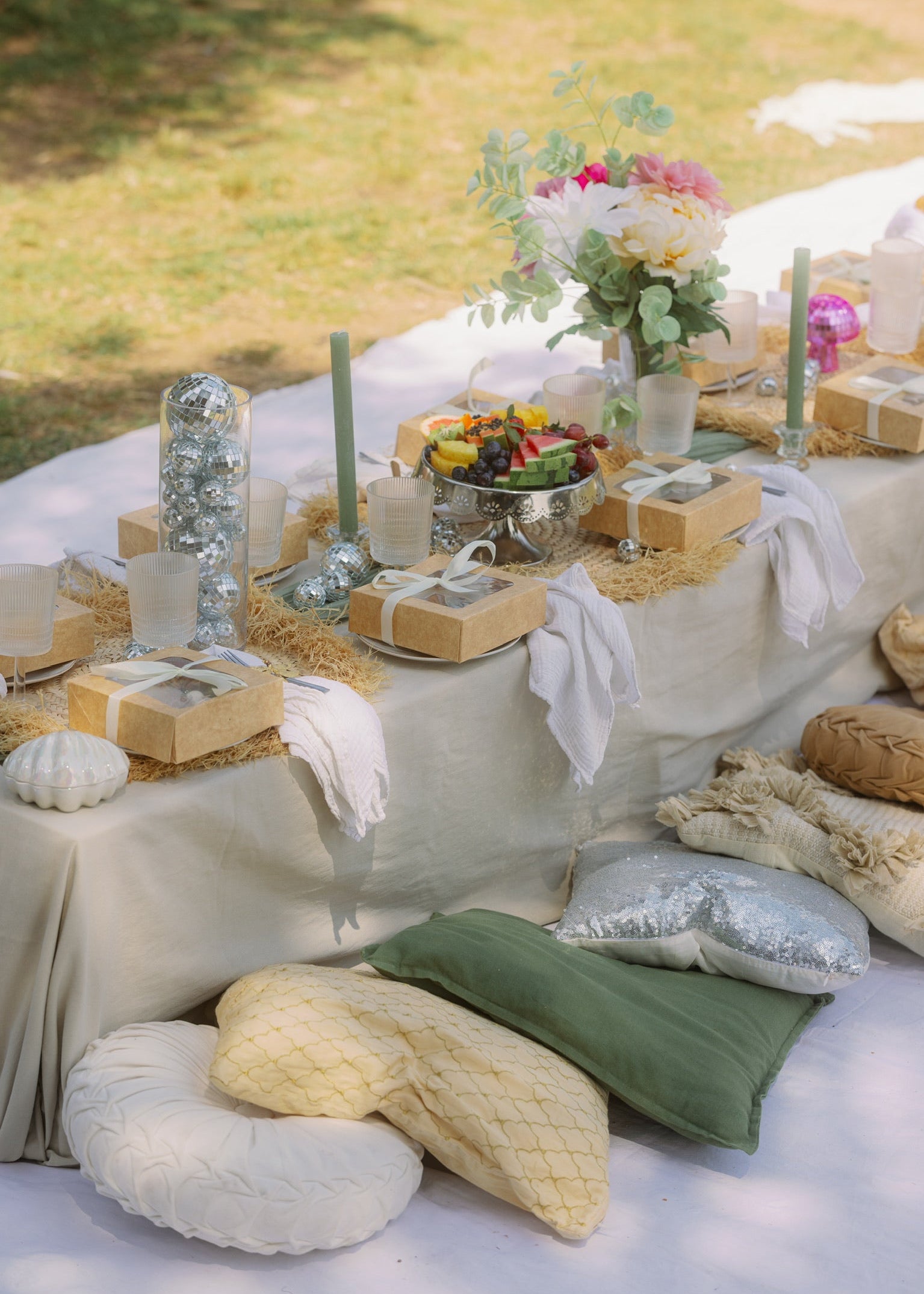 Outdoor setting with a table covered in white cloth, decorated with pillows, flowers, and food items.