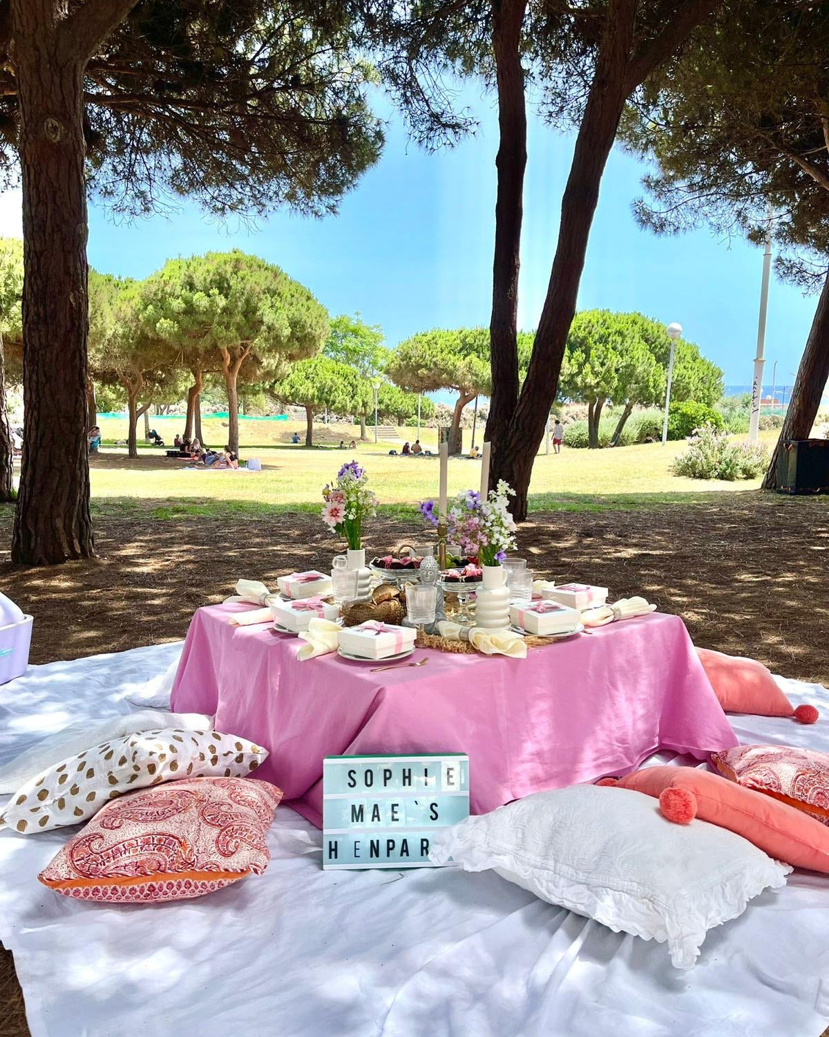 Vibrant hen party picnic in Barcelona  park near the beach, featuring a low table with colorful decorations, fresh flowers, and a view of the sea and palm trees.