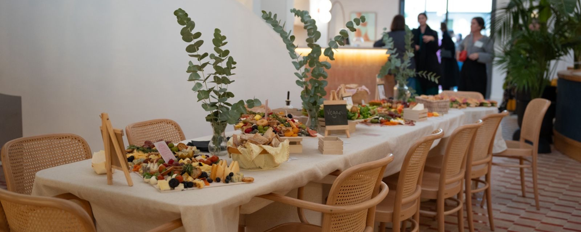 Dining table set for a catering with chairs around, decorated with plants and food.