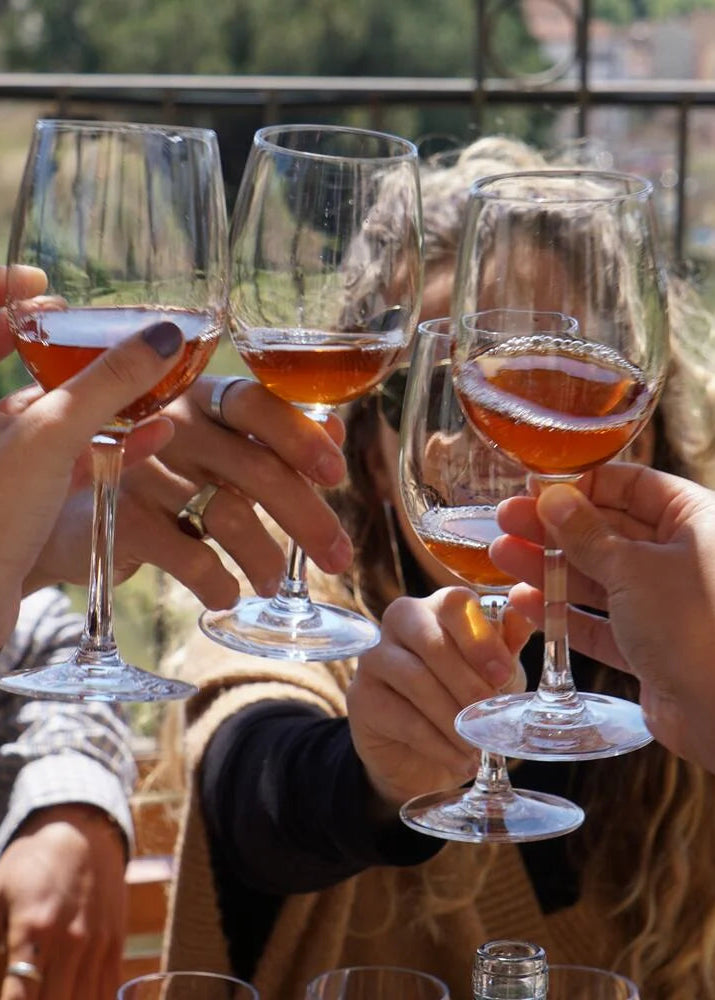 Several people holding up wine glasses filled with an amber-colored beverage in a toast, set against an outdoor vineyard background.