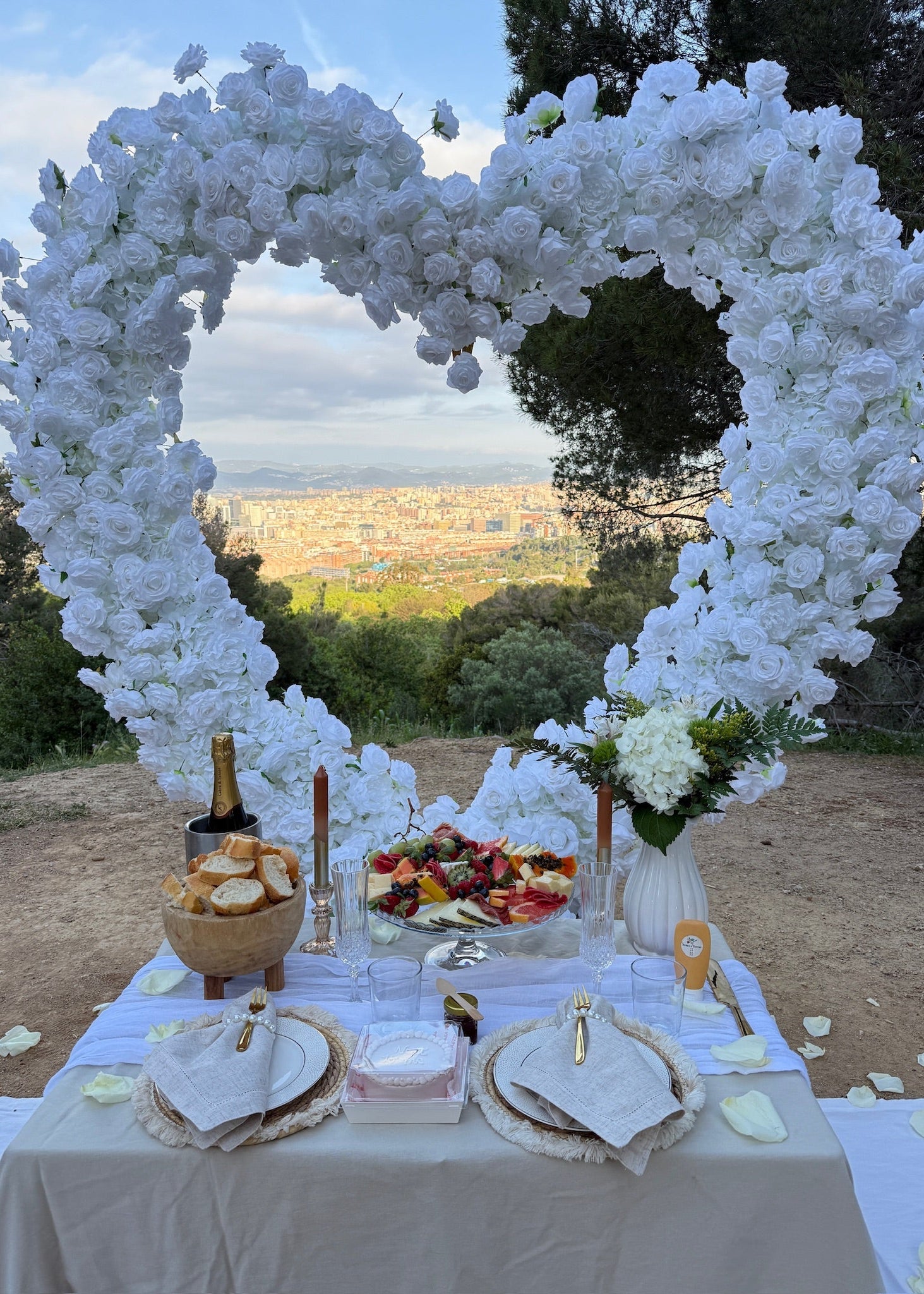 A romantic outdoor wedding-style setup with a large white floral heart-shaped arch over a decorated table. The table is set with white linens, folded napkins, plates, candles, a bottle, bread, fruit, and flowers, with a scenic city view in the background.