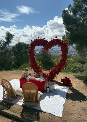 Outdoor romantic setup on a hillside with a large heart-shaped floral arrangement behind a small table covered in a red cloth, two woven chairs, a white blanket, and a scenic city view in the background under a blue sky.