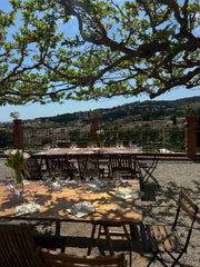 Outdoor dining tables and chairs arranged under a large leafy tree on a terrace, overlooking a scenic hillside landscape with buildings and blue sky.