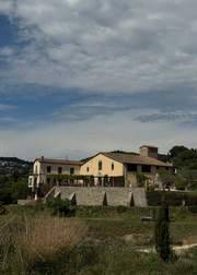 A large rustic villa with a terracotta roof and stone walls, situated in a grassy landscape under a cloudy blue sky.