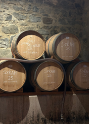 Five wooden barrels stacked on wooden shelves against a rustic stone wall. The barrels are labeled with white chalk text including 'SYRAH 25' and 'APE DI RANELLA'.