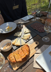 An outdoor dining table set with a charcuterie board featuring sliced bread, cheese wedges, cured meats, crackers, and several wine glasses, with part of a person in a navy sweatshirt visible at the top.