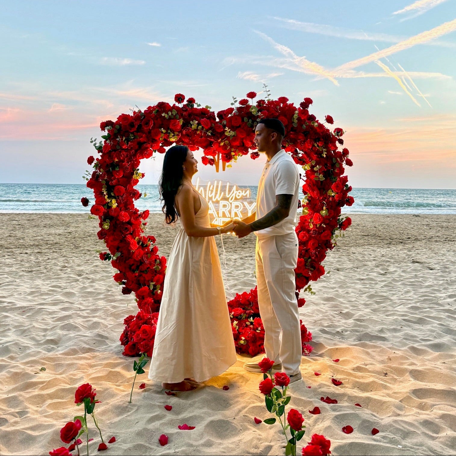 Couple standing under a heart-shaped arch of red flowers on a beach at sunset