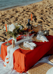 Dining table setup on a sandy beach with red tablecloth, candles, and flowers.