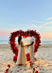 Couple standing under a heart-shaped arch of red flowers on a beach in barcelona for a proposal at sunset.