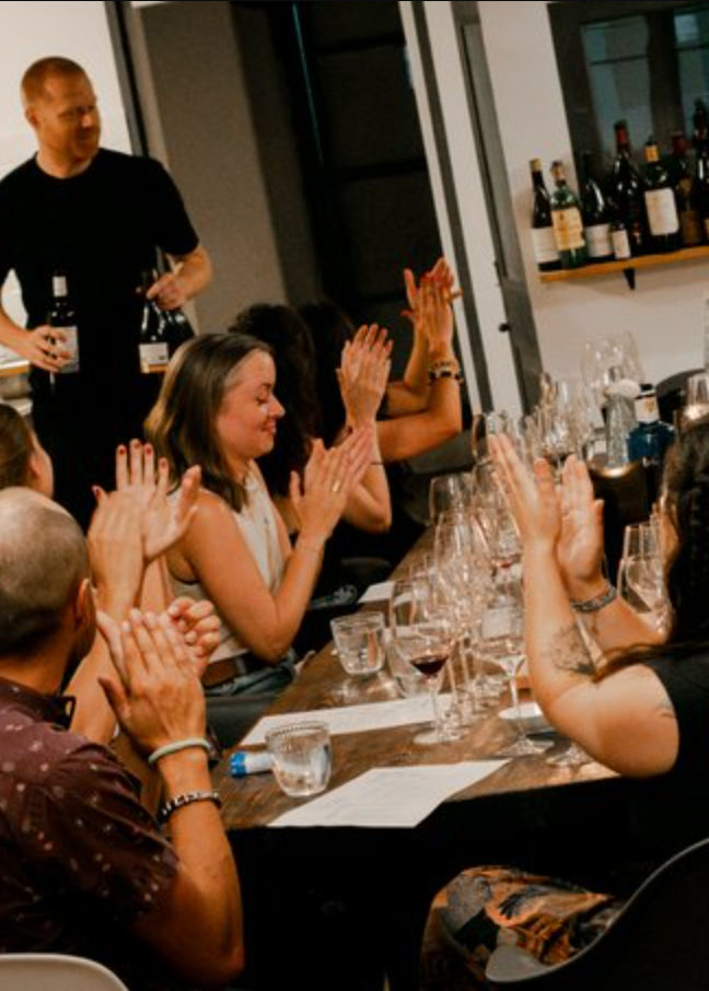 Group of people clapping at a table with wine bottles and glasses in a modern kitchen setting.