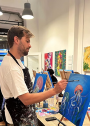 Artist painting a jellyfish on an easel in a studio with other artworks in the background.
