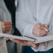 Person holding a notebook and pen with another person in the background