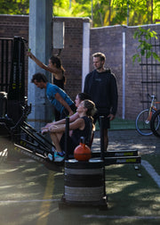 Several people using a Rowing Concord 2 rowing machine in an outdoor backyard gym setting with a brick wall, trees, and a bicycle in the background.