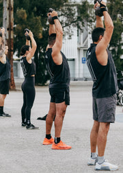 Four people in workout clothes perform a kettlebell overhead press in an outdoor urban plaza. The foreground person wears a black sleeveless top, black shorts, and gray athletic shorts, with orange sneakers.