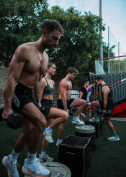 A group of people working out outdoors on a fitness court, with a shirtless man in the foreground lifting a dumbbell while stepping over plyo boxes.
