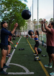 Several people training with medicine balls on an outdoor sports court, with trees, fencing, and other gym equipment visible in the background.