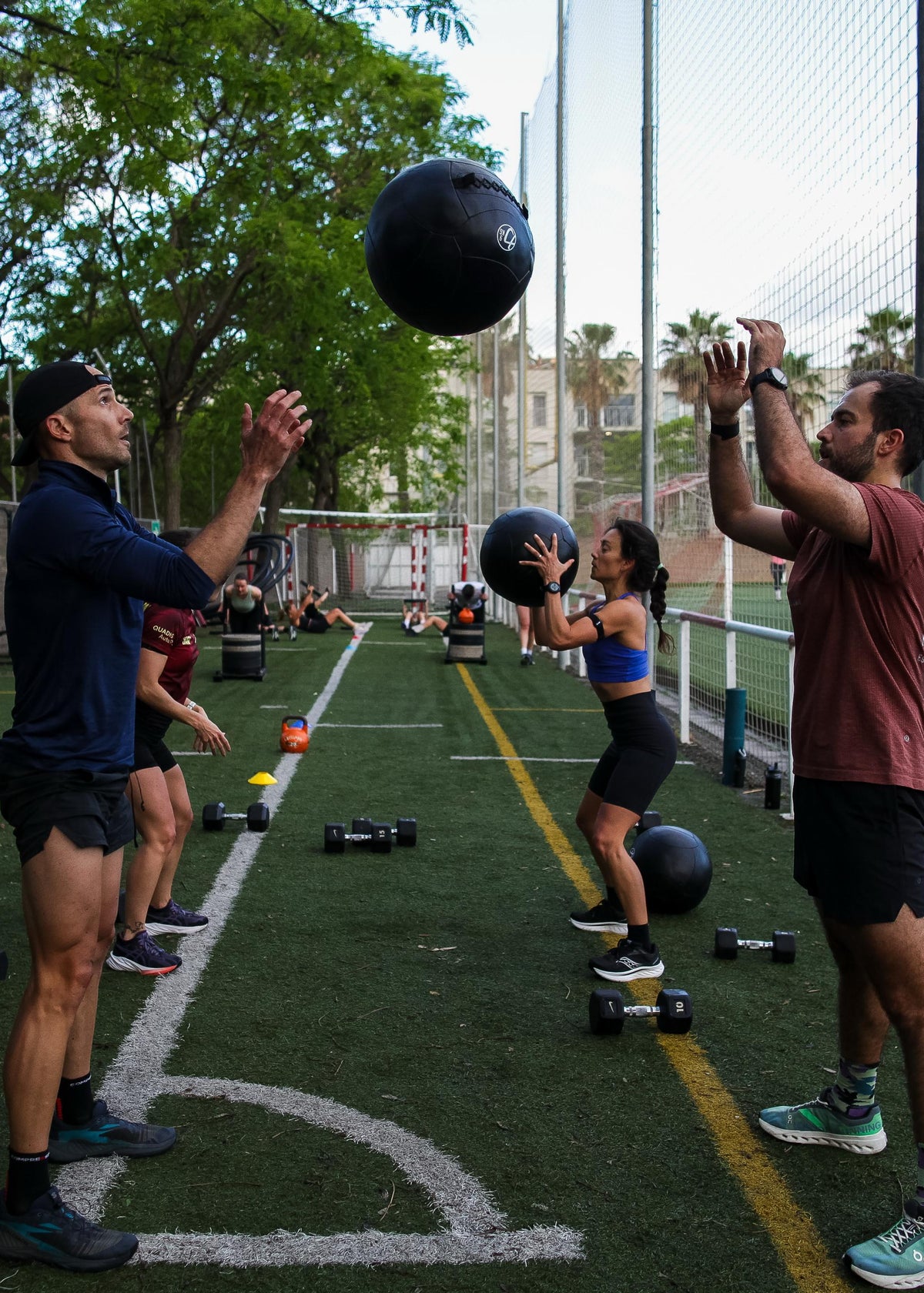 Several people training with medicine balls on an outdoor sports court, with trees, fencing, and other gym equipment visible in the background.