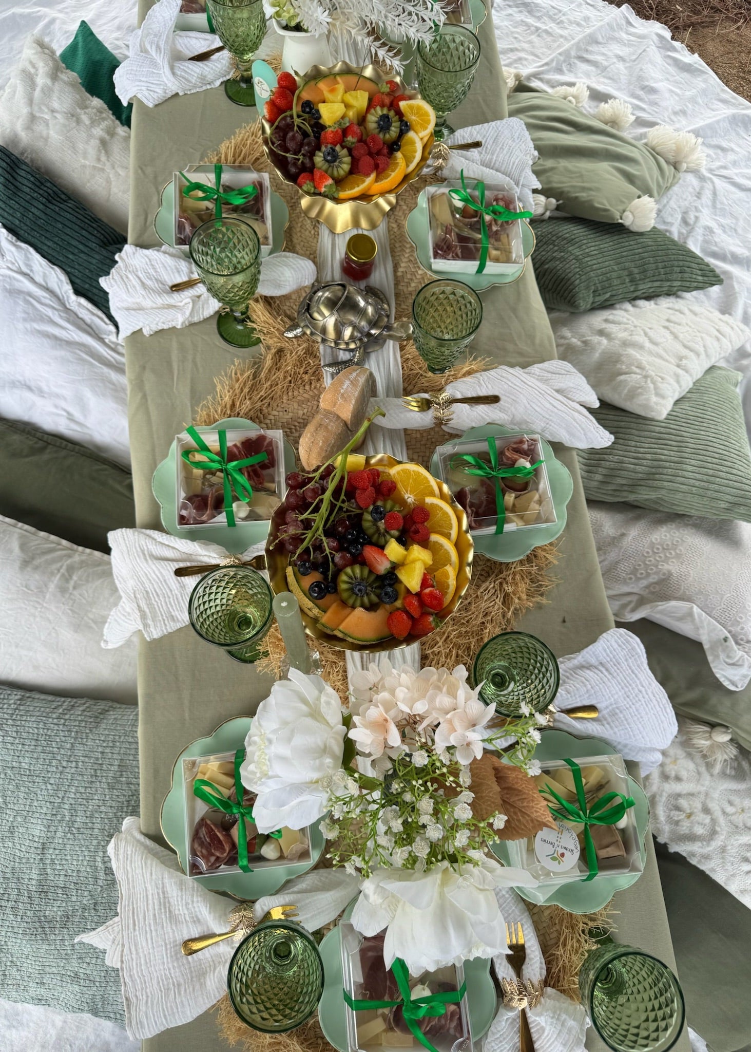 Decorative table setting with fruit, flowers, and green glassware on a bed.