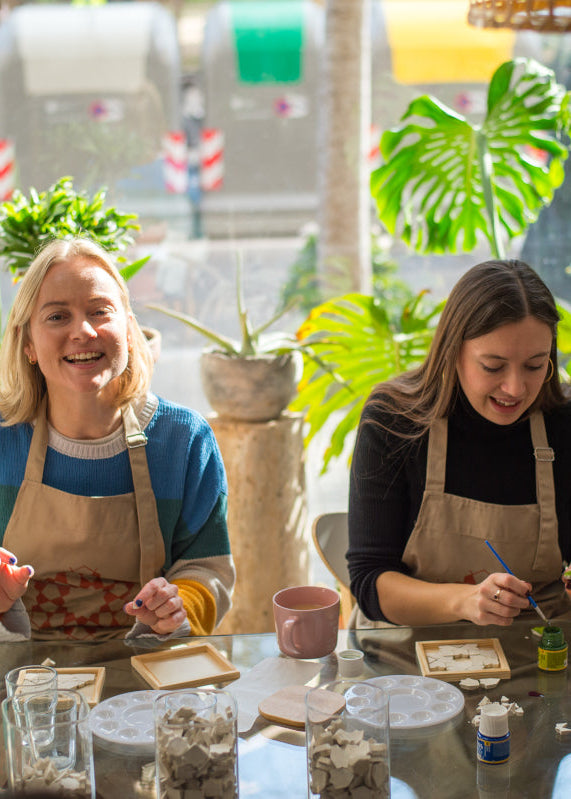 Two women in aprons working at a table with various items in a bright, plant-filled room.