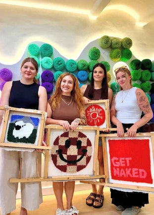 Four women standing together in a brightly lit room, each holding a small framed textile artwork made from colorful yarn. The wall behind them is decorated with many rolled balls of yarn in green, blue, purple, pink, and orange.
