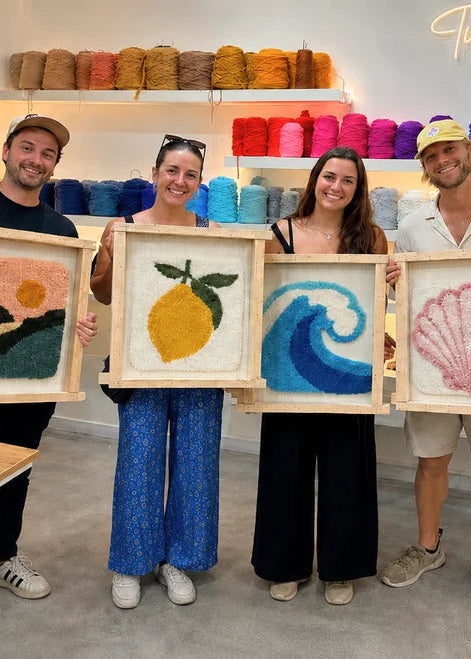 Four people standing in a bright craft studio holding up four framed colorful yarn paintings: a landscape, a lemon, a wave, and a seashell. Shelves in the background are filled with colorful yarn skeins.