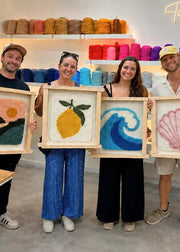 Four people standing in a bright craft studio holding up four framed colorful yarn paintings: a landscape, a lemon, a wave, and a seashell. Shelves in the background are filled with colorful yarn skeins.