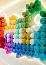 A wall display of many yarn skeins arranged in a rainbow gradient, including red, orange, yellow, green, teal, blue, purple, pink, white, and gray, with warm overhead lighting.