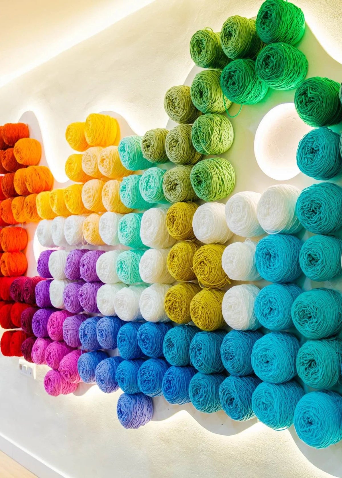 A wall display of many yarn skeins arranged in a rainbow gradient, including red, orange, yellow, green, teal, blue, purple, pink, white, and gray, with warm overhead lighting.