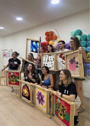 A group of people in a studio holding up framed colorful textile artworks made from yarn, with yarn balls displayed on the wall behind them.